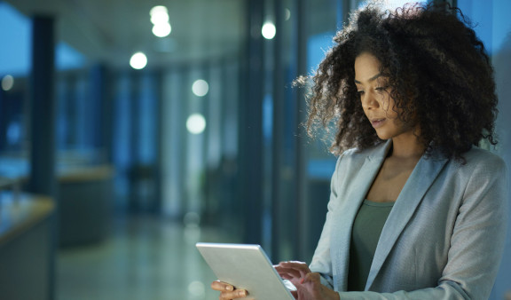 A professional woman with curly hair stands in an office environment during the evening, concentrating on a tablet she's holding. The setting suggests a contemporary workplace with a focus on technology and digital solutions, reflecting the theme of digital consulting.