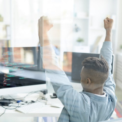 Jeune développeur web levant les mains avec enthousiasme devant son écran d’ordinateur.