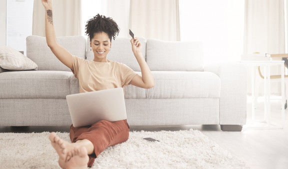 Une jeune femme heureuse et assise par terre dans son salon avec son ordinateur sur les genou surfe sur un site e-commerce