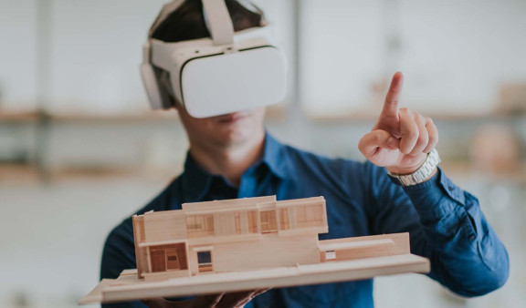 Viewing through VR glasses, a man points on something while holding a wooden model building.