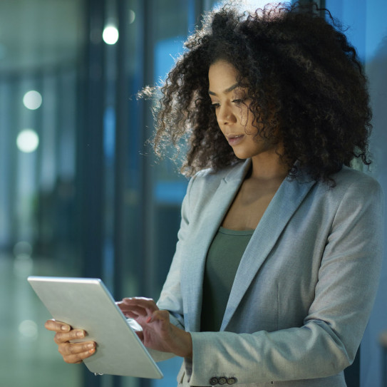A professional woman with curly hair stands in an office environment during the evening, concentrating on a tablet she's holding. The setting suggests a contemporary workplace with a focus on technology and digital solutions, reflecting the theme of digital consulting.